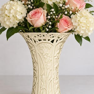 May include: A cream-coloured ceramic vase with an intricate embossed floral pattern and a scalloped, openwork rim. The vase is filled with a lush arrangement of white hydrangea blooms, soft pink roses, and small white baby's breath flowers.