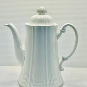 May include: A white porcelain coffee pot with a lid and a curved handle. The pot has a fluted design and a spout for pouring. The lid is topped with a decorative knob. The coffee pot is a classic design.
