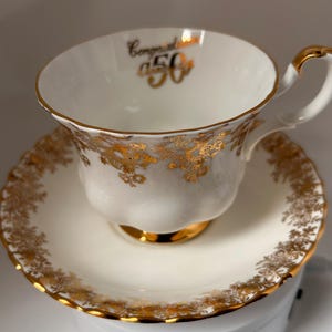 以下が含まれることがあります： 金色の花のディテールとトリムが施された白いティーカップとソーサーのセット。「Congrats 50」の文字が金色の文字で書かれています。カップのハンドルとベースも金色です。