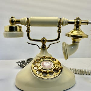 May include: Cream-colored vintage rotary phone with gold-colored accents. The phone features a rotary dial, a handset, and a cradle. The design is reminiscent of classic telephones from the mid-20th century. The phone has a retro aesthetic.