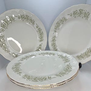 May include: A collection of white ceramic plates with a scalloped edge and a gold trim. Each plate has a green leaf pattern around the rim. The plates are stacked and arranged for display.
