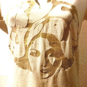 May include: A beige t-shirt with a large, abstract design in shades of brown. The design depicts a stylized face with flowing shapes and lines. The shirt has short sleeves and a crew neck.