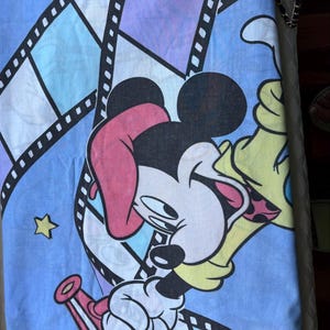 May include: A light blue fabric with a vibrant print of Mickey Mouse and Minnie Mouse. The design incorporates film strip borders, stars, and a megaphone. The characters are depicted in a playful pose, with Minnie wearing a pink bow.