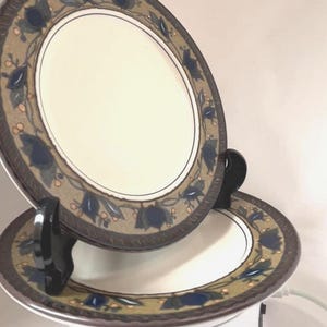 May include: Two round ceramic plates featuring a cream-coloured centre and a wide border decorated with dark blue leaves, small orange berries, and a textured brown outer rim. One plate is displayed upright on a black stand, while the other lies flat.