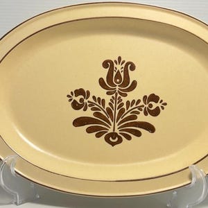 Peut inclure: Un plat de service ovale en céramique de couleur crème claire avec un bord brun foncé. Le centre du plat présente un motif floral brun foncé composé d'une fleur stylisée centrale flanquée de deux fleurs plus petites et de feuillage.