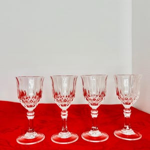 May include: Four clear crystal wine glasses with diamond-cut patterns on a red surface. Each glass has a rounded bowl, a slender stem, and a circular base. The glasses are evenly spaced and arranged in a row.