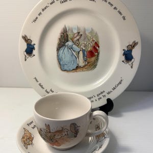 May include: A white ceramic plate, teacup, and saucer set featuring illustrations of rabbits in blue jackets. The plate displays the text: 'Now my dears,' said old Mrs. Rabbit one morning, 'you may go into the fields or down the lane, your father had an accident there; he was put in a pie by Mrs. McGregor.'