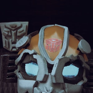 Puede incluir: Primer plano de una máscara de robot gris y naranja con una luz azul que brilla desde los ojos. La máscara tiene un diseño detallado y presenta el símbolo de Autobot en la frente.