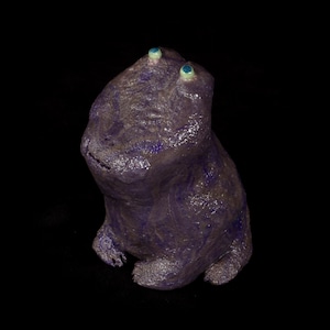 Può includere: Una statuetta di rana in ceramica viola con occhi blu. La rana è seduta eretta con le gambe ripiegate sotto il corpo.