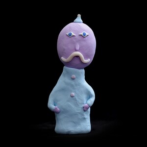 Puede incluir: Una figurita de arcilla de color morado y azul que representa a una persona con bigote y un pequeño sombrero. La figurita lleva una camisa azul claro.