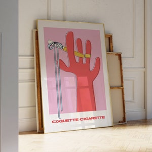 Peut inclure: Une illustration rose et rouge d'une main tenant une cigarette avec un ruban bleu nou&eacute; autour du doigt. Le texte "Coquette Cigarette" est en bas de l'image.