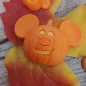 May include: Two orange, 3D-printed plastic decorations shaped like pumpkins with round ears, resembling a cartoon mouse character. The larger pumpkin features a smiling face with visible ridges, while the smaller one is positioned above it.