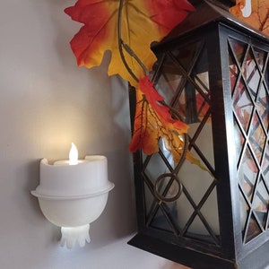 May include: A white candle holder with a flickering flame is mounted on a wall. A black metal lantern with a diamond-shaped pattern is hanging from a vine with fall leaves.