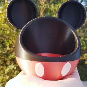 Könnte beinhalten: Ein 3D-gedruckter Behälter im Mickey-Mouse-Design. Der Behälter ist rot mit zwei weißen Ovalen und einem schwarzen Innenraum. Zwei schwarze, kreisförmige Ohren sind oben angebracht. Der Behälter wird in einer Hand gehalten.