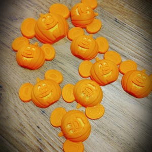 Puede incluir: Un grupo de pequeñas calabazas naranjas con caras de Mickey Mouse talladas en ellas. Las calabazas están sobre una superficie de madera.