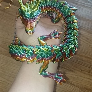 Könnte beinhalten: Eine mehrfarbige, grüne, goldene und rote Drachenfigur mit einem eingerollten Schwanz und detaillierten Schuppen.