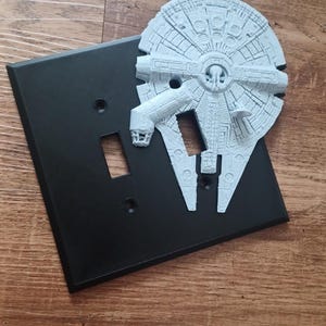 May include: A black light switch cover with a white 3D printed Millennium Falcon from Star Wars.
