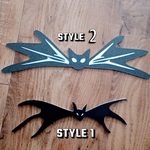 May include: Two black bat silhouettes with white accents. The top bat has a white outline and the bottom bat is solid black. The top bat has the text "STYLE 2" and the bottom bat has the text "STYLE 1".