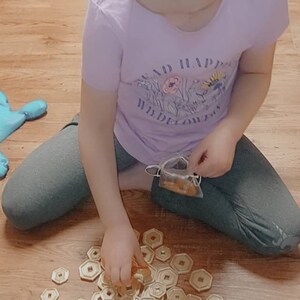 May include: A young person is sitting on a wooden floor, playing with a set of gold-colored, hexagonal-shaped tokens. The tokens are scattered around them, and they are holding one in their hand. The person is wearing a purple shirt with a floral design and the words "Read in wildflowers" printed on it.