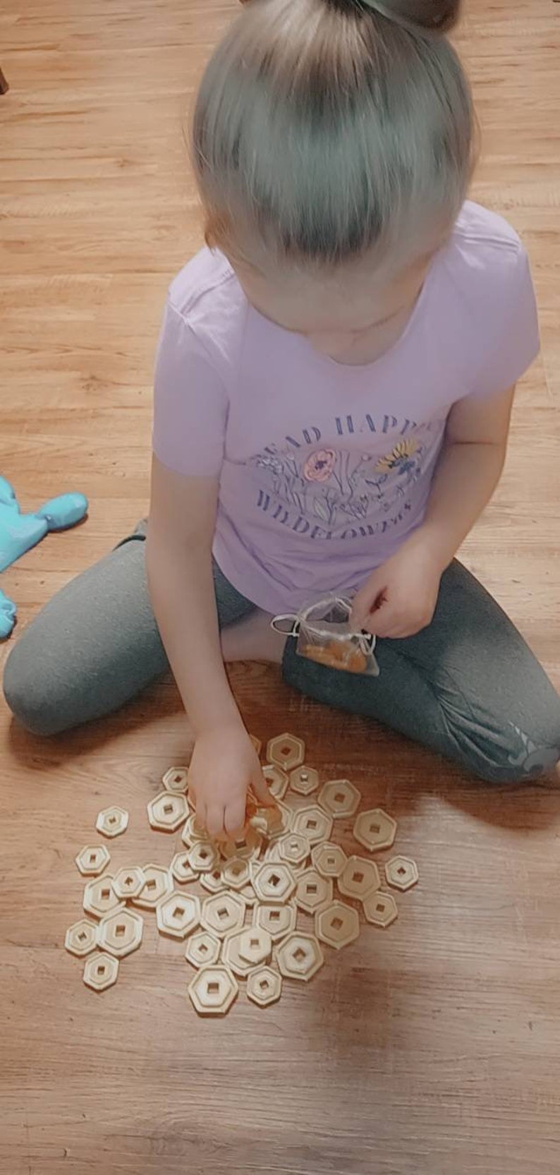 May include: A young person is sitting on a wooden floor, playing with a set of gold-colored, hexagonal-shaped tokens. The tokens are scattered around them, and they are holding one in their hand. The person is wearing a purple shirt with a floral design and the words "Read in wildflowers" printed on it.