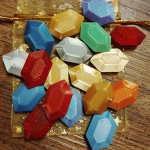 May include: A collection of 15 colorful, glittery, faceted gem-shaped resin charms in a gold mesh bag. The charms are in various shades of blue, green, red, orange, and yellow.