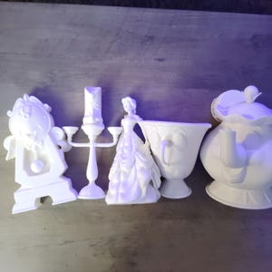 May include: A collection of white ceramic figurines from the Beauty and the Beast movie. The set includes a clock, candelabra, Belle, a teacup, and a teapot. The figurines are arranged on a wooden surface.
