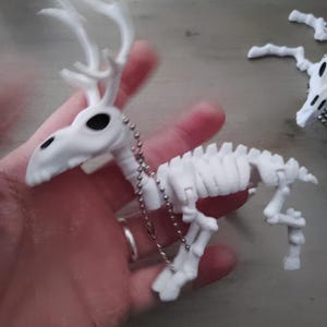 以下が含まれることがあります： 白いプラスチック製の鹿の骨格で、角、黒い眼窩、チェーンが付いています。 骨格のフィギュアは手で持たれており、背景には別の同様のフィギュアが見えます。 ユニークな装飾品です。