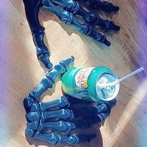 May include: Two black 3D printed skeleton hands holding a green can of soda with a straw. The hands are on a wooden surface.
