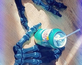 May include: Two black 3D printed skeleton hands holding a green can of soda with a straw. The hands are on a wooden surface.