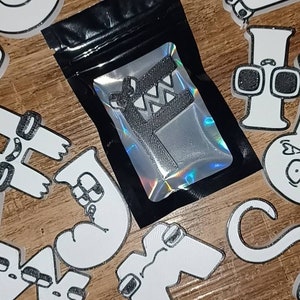 May include: A small black bag with a silver holographic zipper contains a black and white 3D printed object. The bag is surrounded by white 3D printed objects with black outlines, each with a different design.