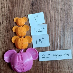 Op de afbeelding: Vier oranje Mickey Mouse-kopvormige magneten in verschillende maten: 2,5 cm, 3,1 cm, 3,8 cm en een roze Mickey Mouse-kopvormige magneet van 6,3 cm.
