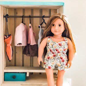 K&ouml;nnte beinhalten: Ein Holz-Puppenhaus-Schrank mit einer Kleiderstange und einer Schublade. Der Schrank ist hellblau gestrichen und hat ein helles Holzinnenleben. Eine Puppe steht vor dem Schrank. Die Puppe tr&auml;gt ein Kleid mit Blumenmuster.