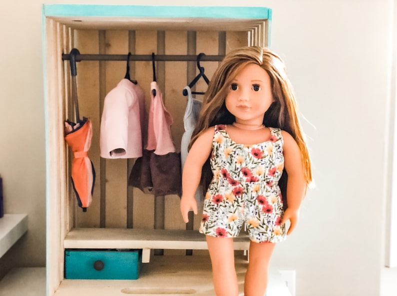 K&ouml;nnte beinhalten: Ein Holz-Puppenhaus-Schrank mit einer Kleiderstange und einer Schublade. Der Schrank ist hellblau gestrichen und hat ein helles Holzinnenleben. Eine Puppe steht vor dem Schrank. Die Puppe tr&auml;gt ein Kleid mit Blumenmuster.