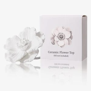 May include: A white ceramic flower top with detailed petals sits next to a white box. The box features a grayscale flower illustration and the text "Ceramic Flower Top (Oil not included)". The flower and box are reflected on a white surface.