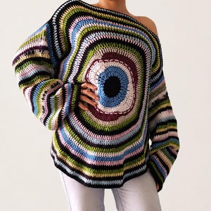 Puede incluir: Un suéter de ganchillo multicolor, con hombros descubiertos. Presenta un diseño circular con anillos concéntricos de varios colores: negro, azul, verde y morado. Las mangas son largas y acampanadas, con un estilo bohemio.