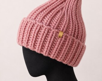 Könnte beinhalten: Ein rosa gerippter Strickbeanie mit einem goldfarbenen Etikett. Der Hut besteht aus einem dicken, strukturierten Garn und hat einen umgeschlagenen Saum. Der Beanie wird auf einem schwarzen Schaufensterkopf präsentiert.