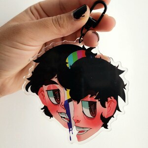 May include: A clear acrylic keychain featuring a split-face illustration of a person with black hair and a colorful stripe design. The keychain has a black clasp for easy attachment.