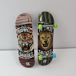 May include: Two miniature skateboards with tiger and lion graphics. The tiger skateboard has the words "PUDWILL SAVAGES" printed on it. The lion skateboard has the words "PUDWILL SAVAGES" printed on it. Both skateboards have black trucks and green wheels.