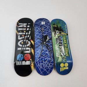 May include: Three fingerboard skateboards with different designs. The first has a red, blue, and white design with the word "Nitro" on it. The second has a blue design with a spider web and a black spider on it. The third has a green, yellow, and blue design with the words "Felipe Tustavo" and "Speed is Key!" on it.