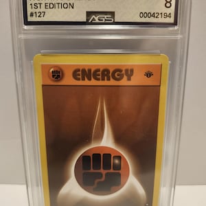 May include: A Fighting Energy trading card from the 2000 P.M. Gym Challenge set, 1st Edition, graded NM-MT 8 by AGS. The card features a stylized energy symbol and the word "ENERGY" in orange and brown tones, encased in a protective case.
