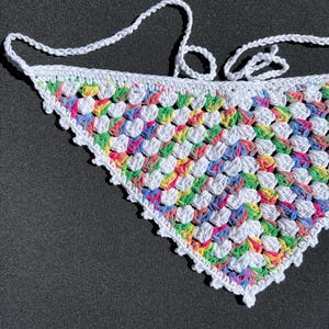 May include: A white crocheted triangle scarf with a colorful, multi-colored pattern. The scarf has a fringe border and is tied with a knot at the top.