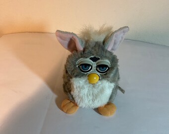 May include: A grey and white furry toy with large blue eyes and a yellow nose. The toy has pink ears and orange feet.
