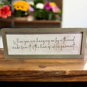 May include: A wooden sign with a light brown background and a dark brown frame. The sign has a white background with black text that reads: "When you are hanging on by a thread, make sure it is the hem of His garment..."