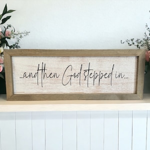 May include: A wooden sign with a white background and a brown frame. The sign says "...and then God stepped in..." in black cursive lettering.