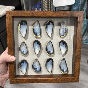 May include: A framed wall art piece featuring twelve blue and white oyster shells with gold accents arranged in a grid pattern on a beige background.