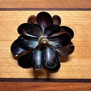 May include: A flower-shaped arrangement of dark purple and blue seashells on a wooden surface.