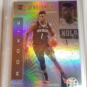 May include: A silver and gold basketball trading card featuring Zion Williamson, a rookie player for the New Orleans Pelicans. The card has a holographic background and the text "Rookie" and "Zion Williamson" in gold lettering.