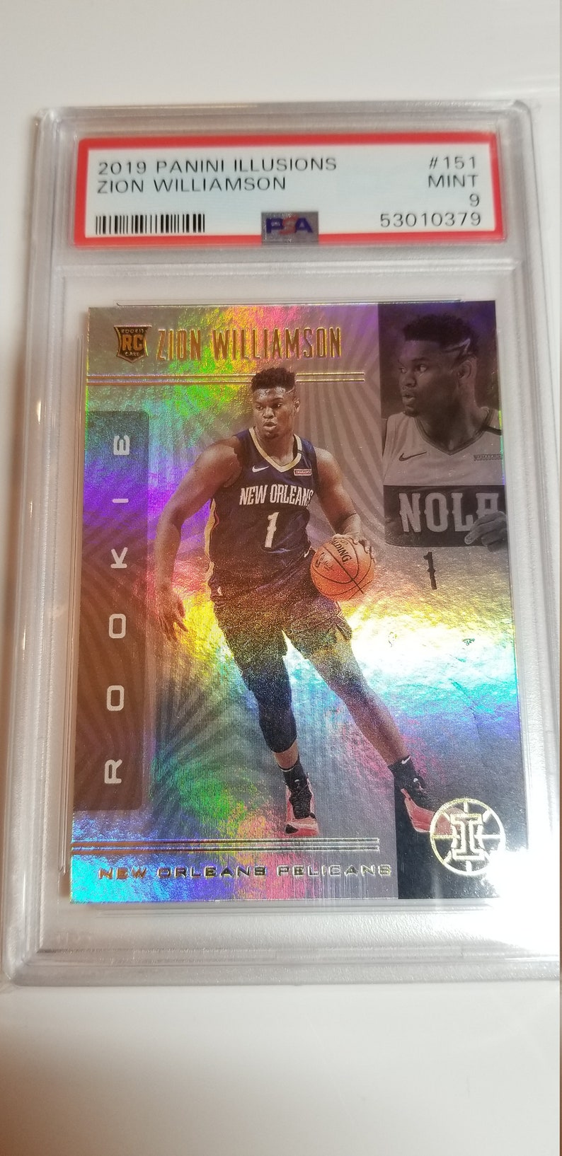 May include: A silver and gold basketball trading card featuring Zion Williamson, a rookie player for the New Orleans Pelicans. The card has a holographic background and the text "Rookie" and "Zion Williamson" in gold lettering.