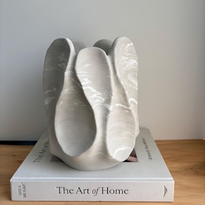 Peut inclure: Une sculpture abstraite gris clair avec des marbrures blanches repose sur un livre intitulé "The Art of Home". La sculpture a une forme unique et organique avec des formes incurvées en forme de pétales. Le livre est gris clair avec du texte noir.