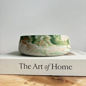 May include: A green and gold marbled concrete bowl with a white background. The bowl is sitting on a book titled "The Art of Home".